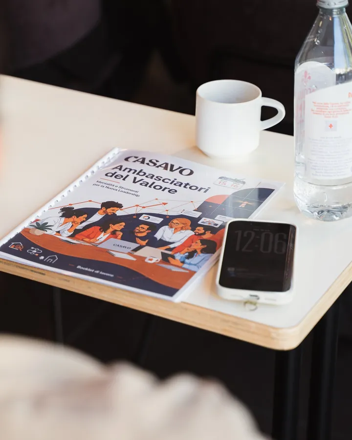 Il booklet di lavoro Casavo «Ambasciatori del Valore — Mentalità e strumenti per la nuova leadership» su un banco, accanto a una tazza e a uno smartphone.